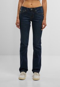 Model îmbrăcat în blugi bleumarin și adidași albi, stând în fața unui fundal gri deschis. Topul ajustat evidențiază abdomenul.