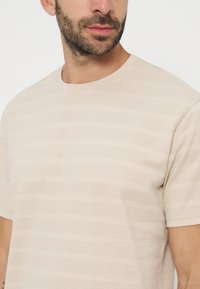 Homme avec une barbe courte portant une chemise beige texturée à manches courtes avec des rayures horizontales, regardant légèrement sur le côté.