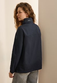 Navyblauwe lichtgewicht jas met een hoge kraag, elastische manchetten en subtiele branding op de mouw. Soepele textuur en een relaxte pasvorm.