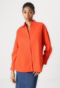 Camicia button-up arancione brillante con colletto, vestibilità rilassata, maniche lunghe e una texture liscia, abbinata a una gonna di jeans.