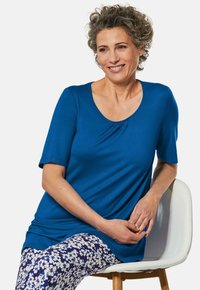 Blaues, kurzärmeliges Oberteil mit einem runden Ausschnitt und gerafftem Detail. Kombiniert mit floral gemusterten Leggings, sitzt auf einem weißen Stuhl.