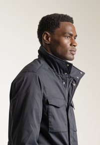 Chaqueta negra impermeable con collar alto, cierre de cremallera y dos bolsillos en el pecho. Textura suave con cierres a presión y ajuste entallado.