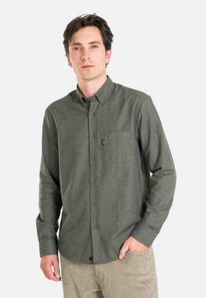 Mann mit dunklem Haar, der ein grünes Hemd mit Knöpfen und Tasche sowie beige Cordhosen trägt, steht mit einer Hand in der Tasche.