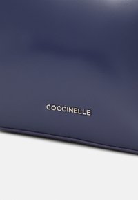 Borsa a mano in pelle blu navy con una texture liscia, caratterizzata da un logo del brand "Coccinelle" in argento impresso sulla parte anteriore.