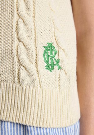 Cream cable-knit sweater vest with green embroidered logo above ribbed hem, worn over blue and white striped pants.