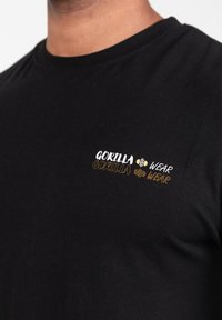Juodas medvilninis marškinėliai su daugiamačiu "GORILLA" teksto logotipu kairėje krūtinės pusėje, papuoštas baltais ir auksiniais akcentais. Lygi tekstūra.