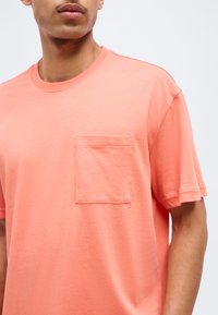 Άνδρας που φοράει κοραλλί κοντομάνικη μπλούζα με τσέπη στο στήθος, εμφανιζόμενος από τον λαιμό έως τη μέση, με φόντο απλό.