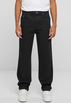 Man draagt losse zwarte jeans, witte sneakers en een wit t-shirt dat in de broek is gestopt, staand tegen een effen grijze achtergrond.