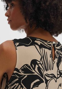Sleeveless blouse in beige with bold black floral pattern, featuring a small keyhole and button closure at the back. Smooth, lightweight fabric.