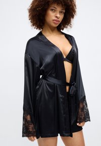 Čierny saténový kimono s dlhými, voľnými rukávmi, čipkovaným lemom na manžetách a viazaným opaskom v páse, nosené cez zodpovedajúci čierny spodný prádlo.