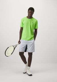 T-shirt sportivo verde neon, tessuto leggero; abbinato a pantaloni corti grigi chiari con una striscia laterale nera; in mano una racchetta da tennis.