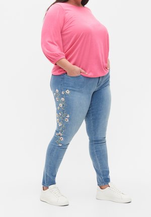 Femme portant un haut rose à manches longues, un jean bleu clair avec une broderie florale blanche sur la cuisse gauche, et des baskets blanches, mains dans les poches.
