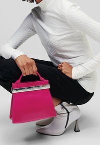 Różowa torebka z srebrnymi akcentami, o zorganizowanej konstrukcji i krótkich uchwytach. Modelka nosi teksturowany biały top i czarne spodnie, zestawione z białymi botkami sięgającymi kostek.