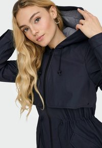 Veste à capuche marine avec taille froncée, cordons ajustables et doublure rayée à l'intérieur de la capuche. Dispose d'une fermeture éclair et de poches.