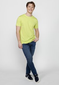 Tricou scurt cu mânecă scurtă de culoare verde deschis, cu decolteu rotund și un logo subtil. Asortat cu blugi albaștri și adidași negri, într-o poziție relaxată.