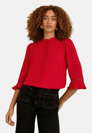 Rode blouse met een gekartelde halslijn, geplooide voorkant en uitlopende mouwen met decoratieve stiksels. Draag het met zwarte jeans.