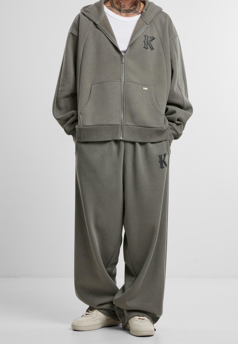 Personne portant un sweat à capuche zippé oversize vert olive et un pantalon de survêtement large assorti avec des logos noirs en forme de "K", accompagné de baskets blanches.