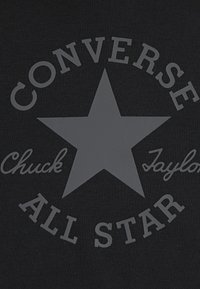 Material negru cu o stea mare gri și textul „CONVERSE” deasupra și „ALL STAR” dedesubt, cu „Chuck Taylor” într-un font script.