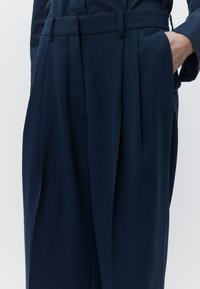 Calças largas azul-marinho com cintura alta, pregas, passante para cinto e bolsos laterais. Tecido macio com textura suave.