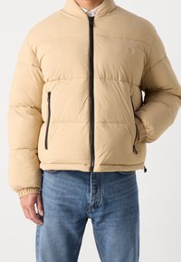 Beige puffer-takki, jossa on vetoketjullinen etuosa, sivutaskut, elastiset hihansuut ja sävy sävyyn -logo. Takissa on laatikkomainen, pehmustettu muotoilu ja sileä pinta.