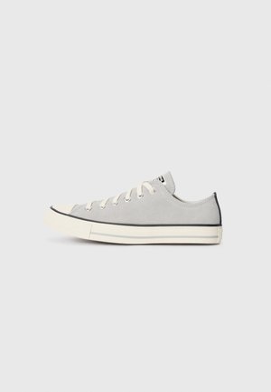 Jasnoszary niski sneaker z białą gumową podeszwą, białymi sznurowadłami i czarnymi lamówkami, pokazany z profilu na jednolitym jasnym tle.