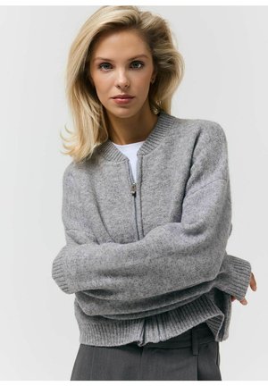 Blondynka w szarym swetrze rozpinanym na zamek, na białej koszuli, ze skrzyżowanymi ramionami, pozuje na tle jednolitego jasnoszarego tła.