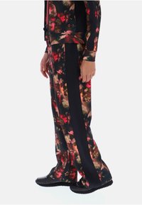 Persona che indossa pantaloni a fantasia floreale con pannelli laterali neri e sneaker nere, in piedi su uno sfondo bianco.