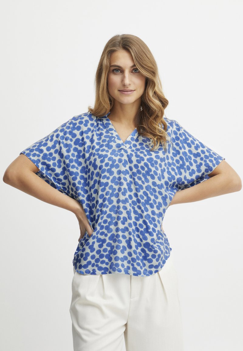 Top blu con maniche corte e scollo a V, decorato con un motivo di punti blu di varie dimensioni su uno sfondo azzurro chiaro. Tessuto leggero.