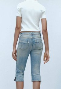 Pantalones capri de mezclilla azul con acabado desteñido, con bolsillos traseros y un dobladillo que llega justo por debajo de la rodilla. Combinados con una camiseta blanca de manga corta.