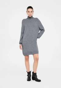 Grijze gebreide sweaterjurk met een hoge hals, lange mouwen en een losse pasvorm, gecombineerd met zwarte enkellaarsjes met gespsdetails.