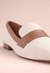 Mocassin en toile beige avec une lanière en cuir marron sur le dessus. Présente un talon bas et un design à bout pointu. Matière texturée.