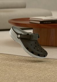 Black and gray Croc-style shoe floating above a beige textured carpet with a wooden table and sofa in the background.