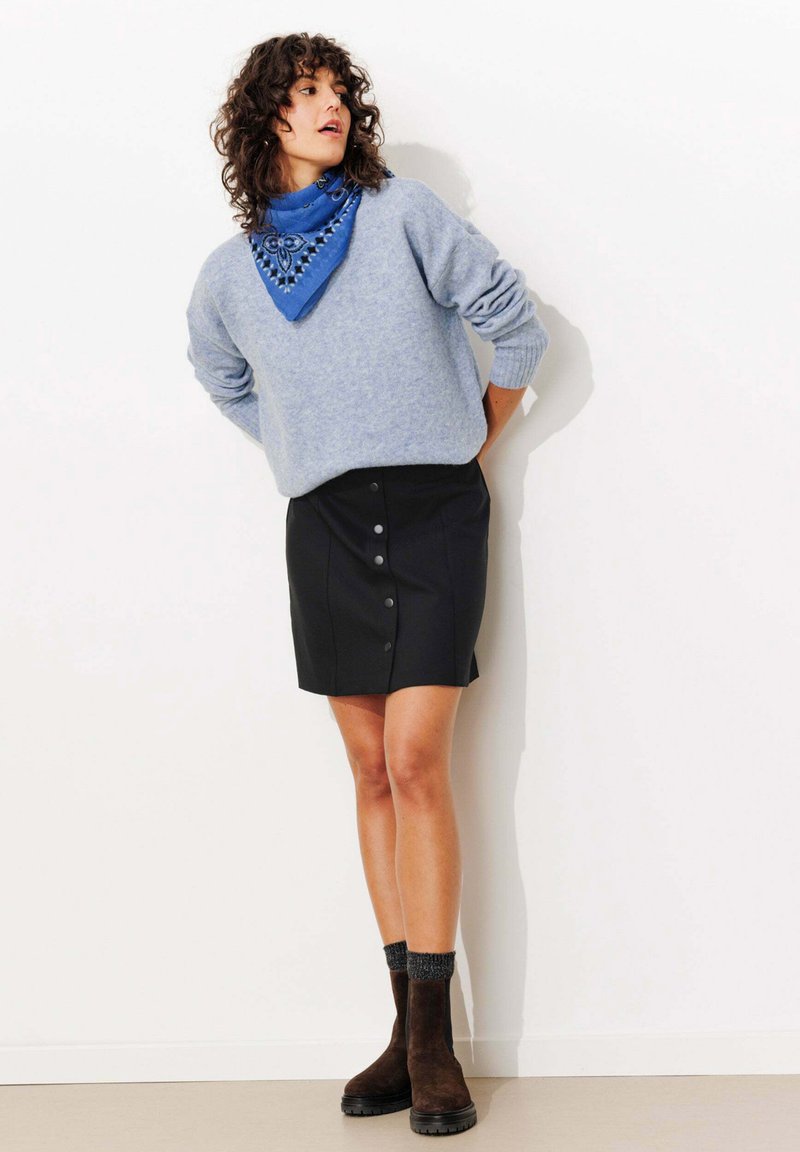 Femme portant un pull bleu clair, une écharpe de cou à motifs bleus, une mini-jupe noire à boutons, des chaussettes foncées et des bottines brunes, se tenant devant un mur blanc.