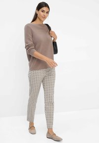 Pull en tricot marron clair à manches longues, associé à un pantalon à motifs beige et gris. Le mannequin porte des ballerines grises et tient un sac noir.