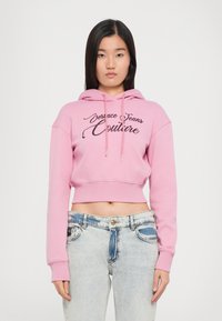 Felpa rosa corta con cappuccio regolabile, caratterizzata da testo ricamato nero "Versace Jeans Couture". Indossata con jeans in denim chiaro.