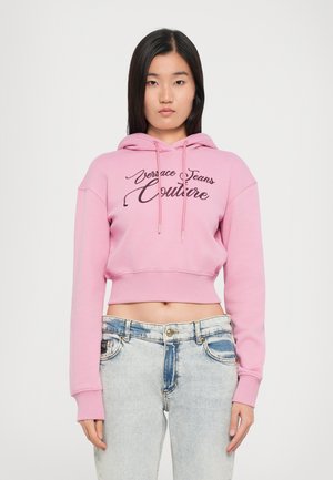 Rosa cropped hettegenser med snorhette, med svart brodert "Versace Jeans Couture"-tekst. Brukt med lyse denimjeans.