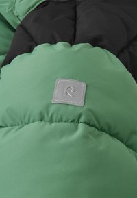 Sezione di giacca a vento verde con design trapuntato, caratterizzata da una sezione nera e una toppa con logo in tessuto grigio sul davanti. Texture morbida e liscia.