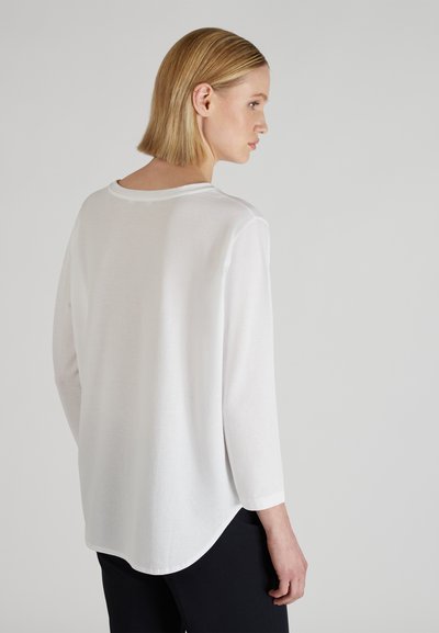 Biała bluzka z długim rękawem, okrągłym dekoltem i zaokrąglonym dołem, wykonana z lekkiego materiału, o luźnym kroju i prostym designie.