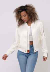 Veste bomber crème avec fermeture éclair à l'avant, poignets côtelés et poches latérales. Présente une broderie discrète et une étiquette orange à l'ourlet.