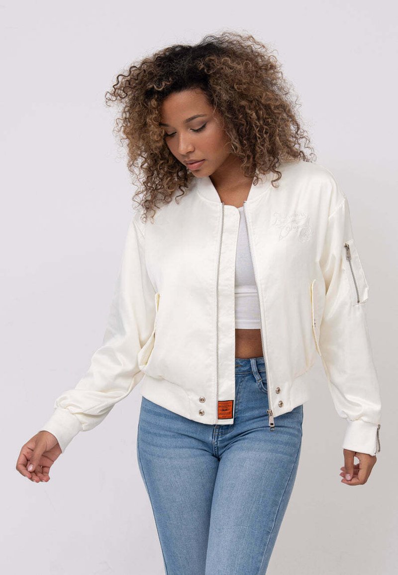 Veste bomber crème avec fermeture éclair à l'avant, poignets côtelés et poches latérales. Présente une broderie discrète et une étiquette orange à l'ourlet.