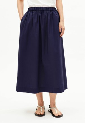 Femme portant une jupe midi bleu marine avec une taille élastique et des sandales blanches tressées, debout les mains dans les poches.