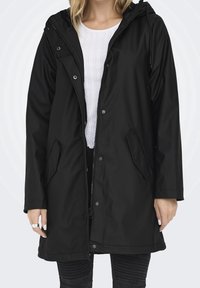 Femme portant un long imperméable noir à capuche par-dessus une chemise blanche texturée et un pantalon noir côtelé, debout devant un fond uni.