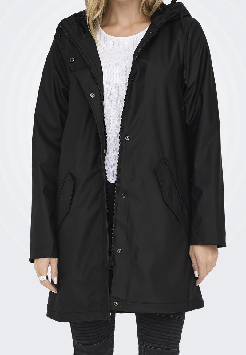 Femme portant un long imperméable noir à capuche par-dessus une chemise blanche texturée et un pantalon noir côtelé, debout devant un fond uni.