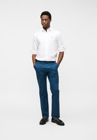 Νεαρός άνδρας στέκεται με τα χέρια στις τσέπες μπλε παντελονιών, φορώντας λευκό πουκάμισο με κουμπιά και μαύρα μοκασίνια, σε απλό φόντο.