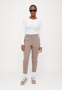 Vit långärmad topp, beige skräddarsydda byxor med uppvikt kant och beige sneakers med grå detaljer. Modellen bär svarta solglasögon.