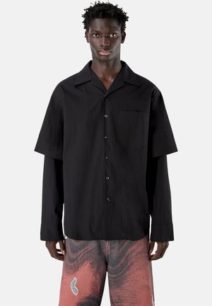 Donkere huidkleurige persoon die een gelaagd zwart overhemd draagt met korte en lange mouwen, en roze broek met patroon, tegen een effen achtergrond.