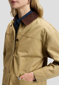 Femme portant une veste beige avec un col en velours côtelé marron et une chemise bleue, main dans la poche de la veste, montrée du cou à la taille.