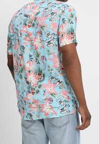 Chemise à manches courtes en tissu bleu clair, ornée de motifs floraux vifs en rose, vert et noir. Coupe décontractée, texture douce.
