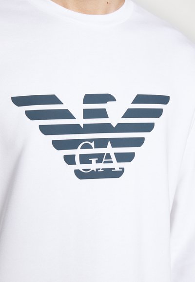 T-shirt blanc à manches longues présentant un logo d'aigle bleu foncé avec des rayures horizontales et les lettres "GA" centrées en dessous de l'aigle.