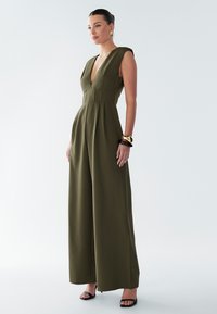 Combinaison verte olive avec un décolleté en V plongeant, une coupe à jambes larges et un corsage structuré. Tissu lisse avec de minimalistes accents et des sandales noires épurées.